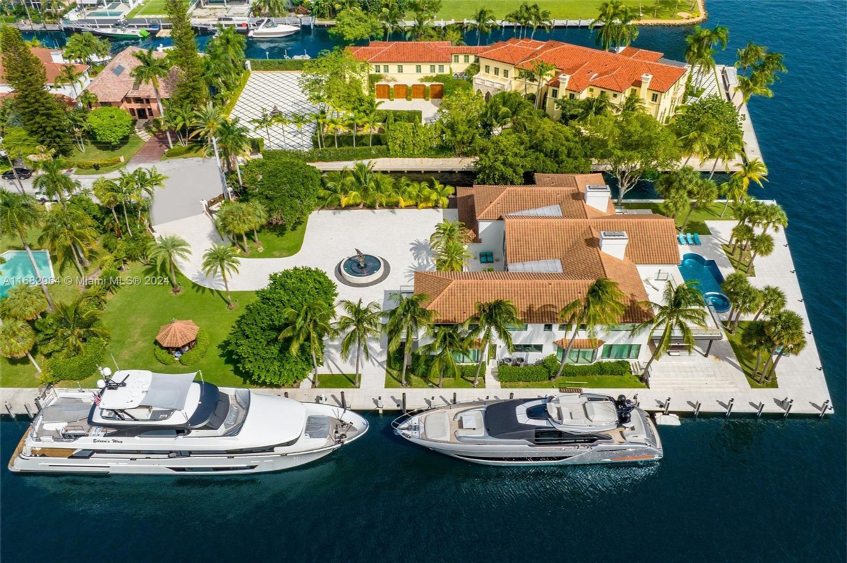 Aerial view of a luxurious waterfront estate showcasing sprawling lawns, palm-lined driveways, and two large yachts docked along the private marina.