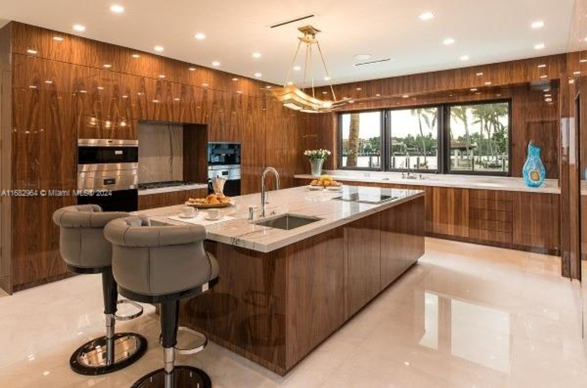 Kitchen showcases high-gloss wood cabinetry, sleek appliances, and a large marble island with waterfront views.