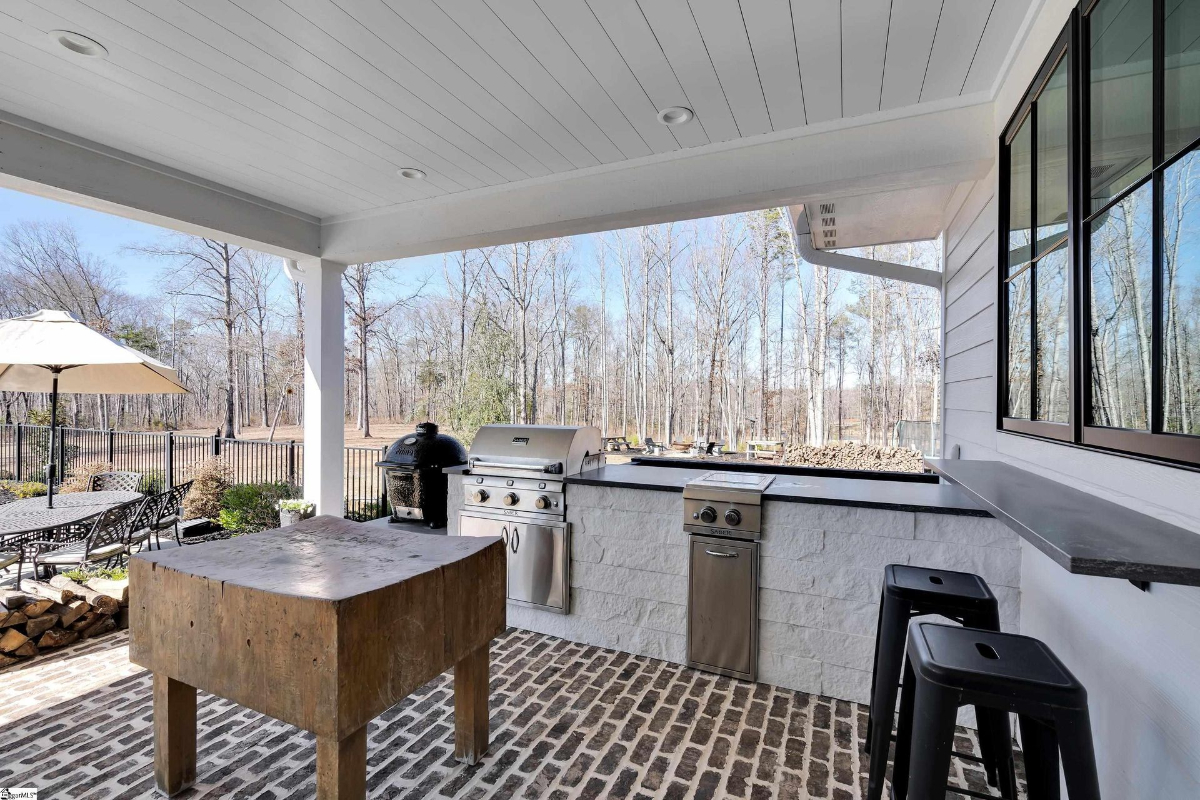 Outdoor kitchen equipped with premium grills, a butcher block table, and bar seating overlooking a wooded landscape.