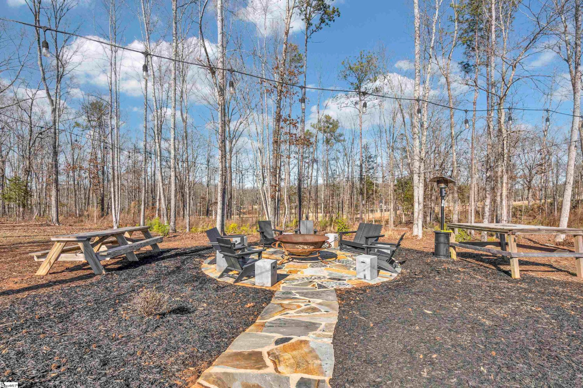 Cozy outdoor fire pit area surrounded by Adirondack chairs, picnic tables, and string lights nestled in a wooded setting.