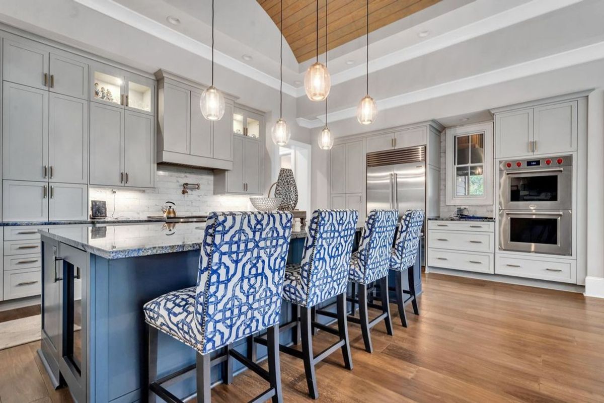 A gourmet kitchen boasts a striking island with patterned blue chairs under a vaulted wooden ceiling.