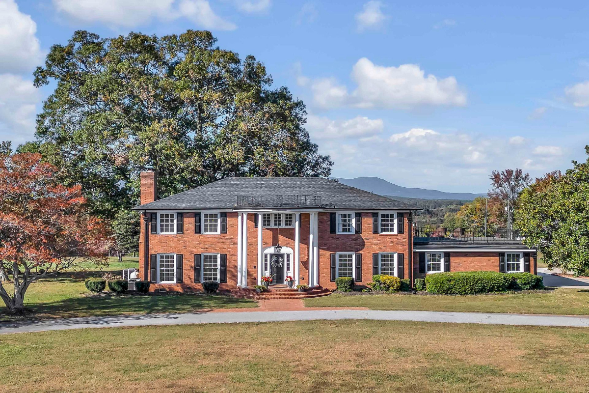  Elegant colonial-style brick home with classic white columns, surrounded by lush greenery and mountain views in the background.