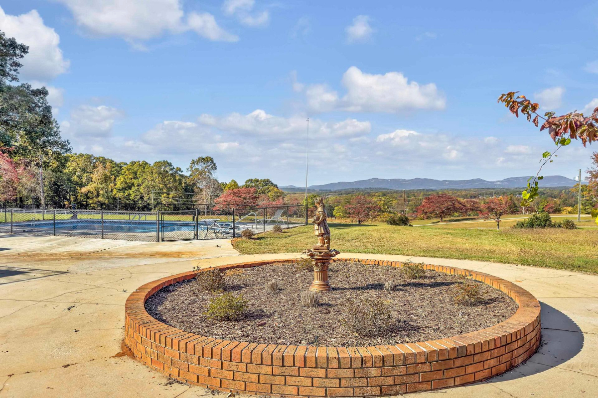 A charming brick-bordered flower bed with a decorative statue is set against a backdrop of a fenced pool and rolling hills in the distance.