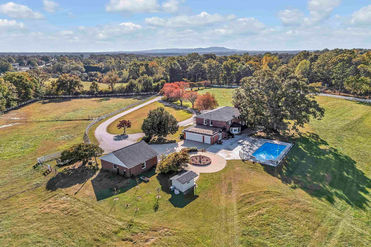 Aerial view of a picturesque estate with a circular driveway, expansive pastures, a pool, and additional outbuildings, all surrounded by lush greenery.