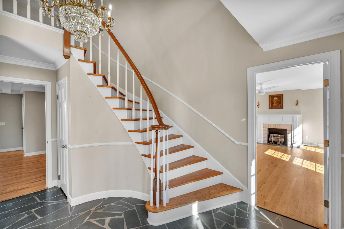 The foyer showcases a timeless curved staircase with warm wood tones, slate flooring, and access to a sunlit living room with a classic fireplace.