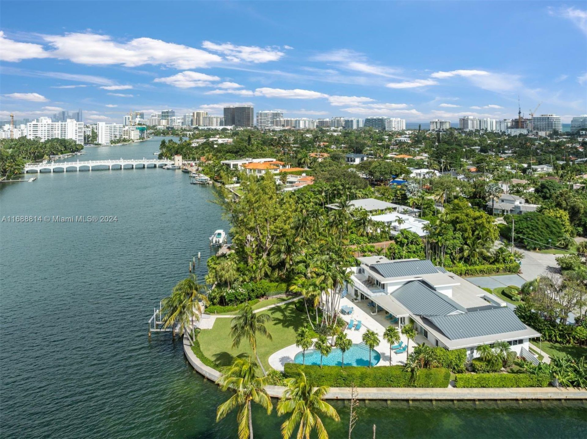 Aerial view of a waterfront estate featuring a private dock, lush landscaping, and a serene pool area, with cityscape in the background.