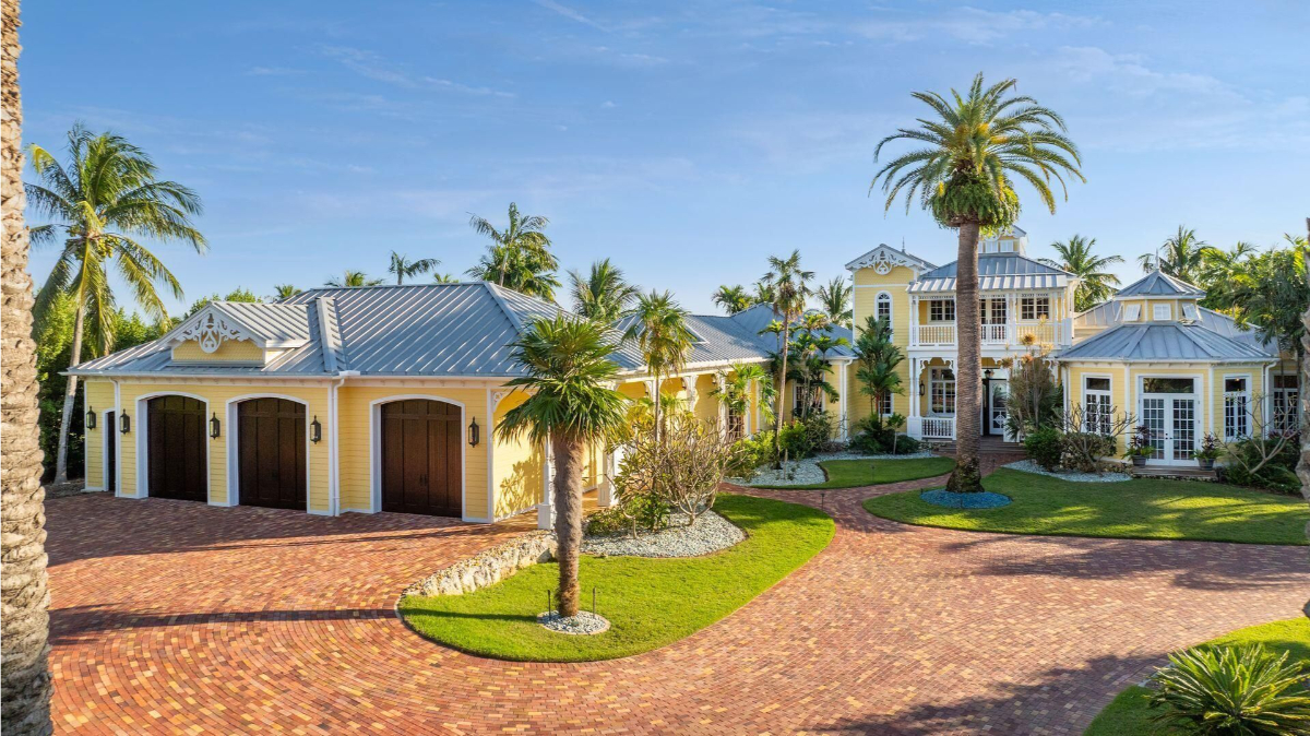 A vibrant coastal estate features cheerful yellow siding, a metal roof, lush tropical landscaping, and a classic brick driveway leading to a three-car garage.