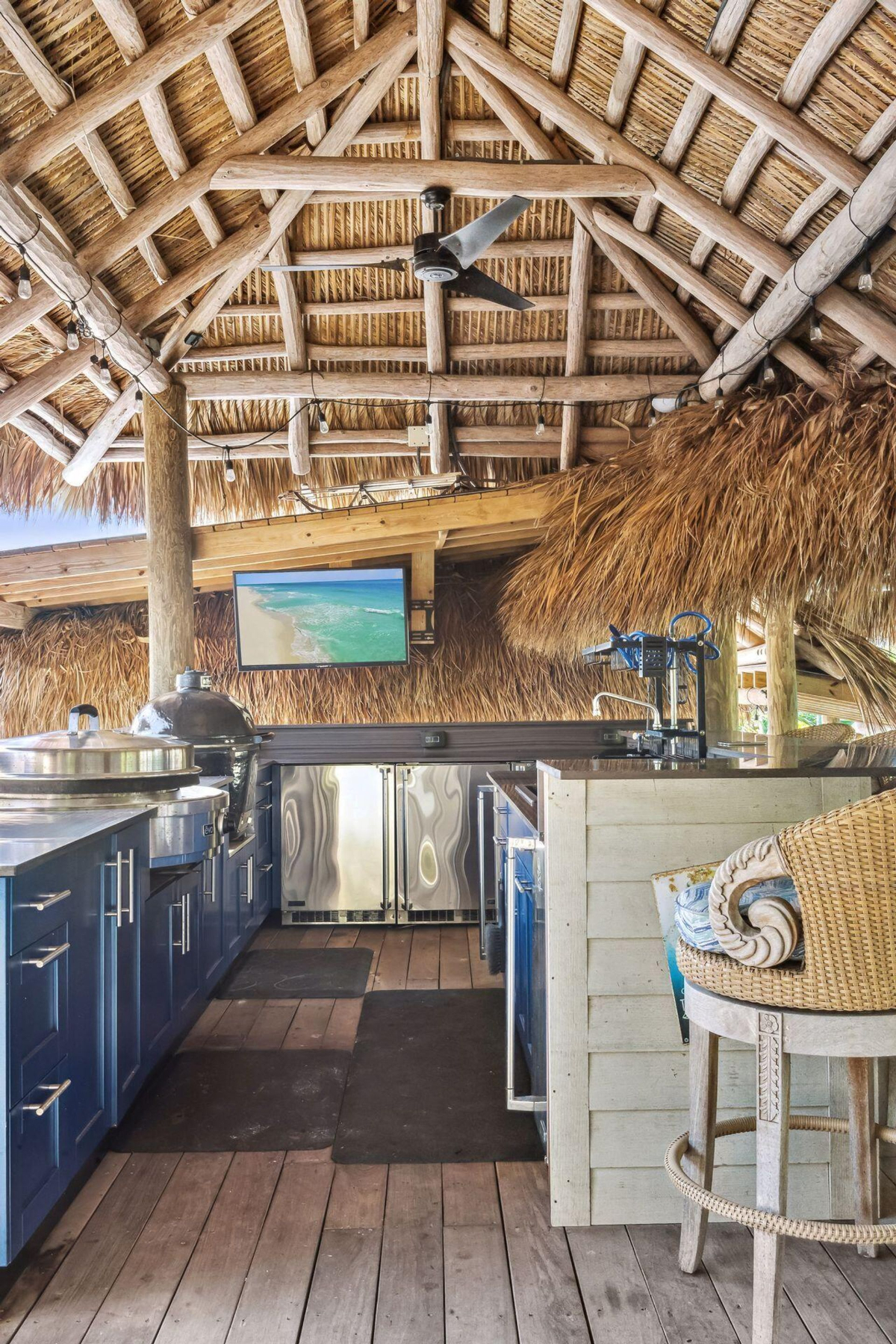 Outdoor kitchen under a tiki hut features stainless steel appliances and a flat-screen TV, ideal for entertaining.