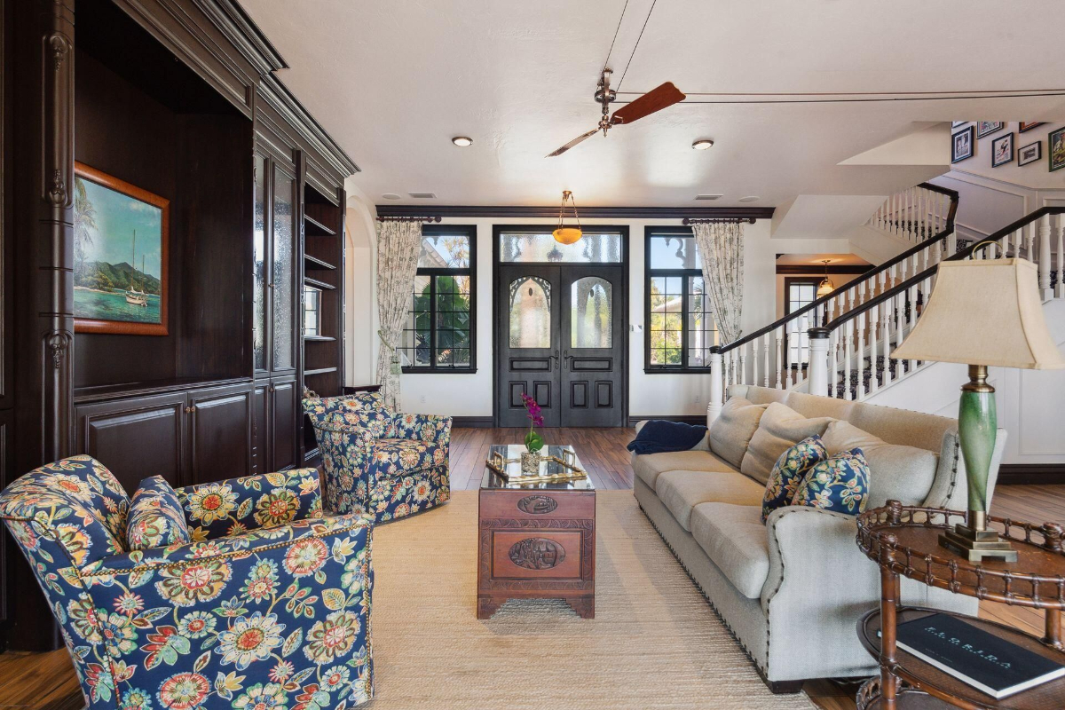 A welcoming living room features floral-patterned armchairs, dark wood cabinetry, and a central view of double front doors framed by large windows.