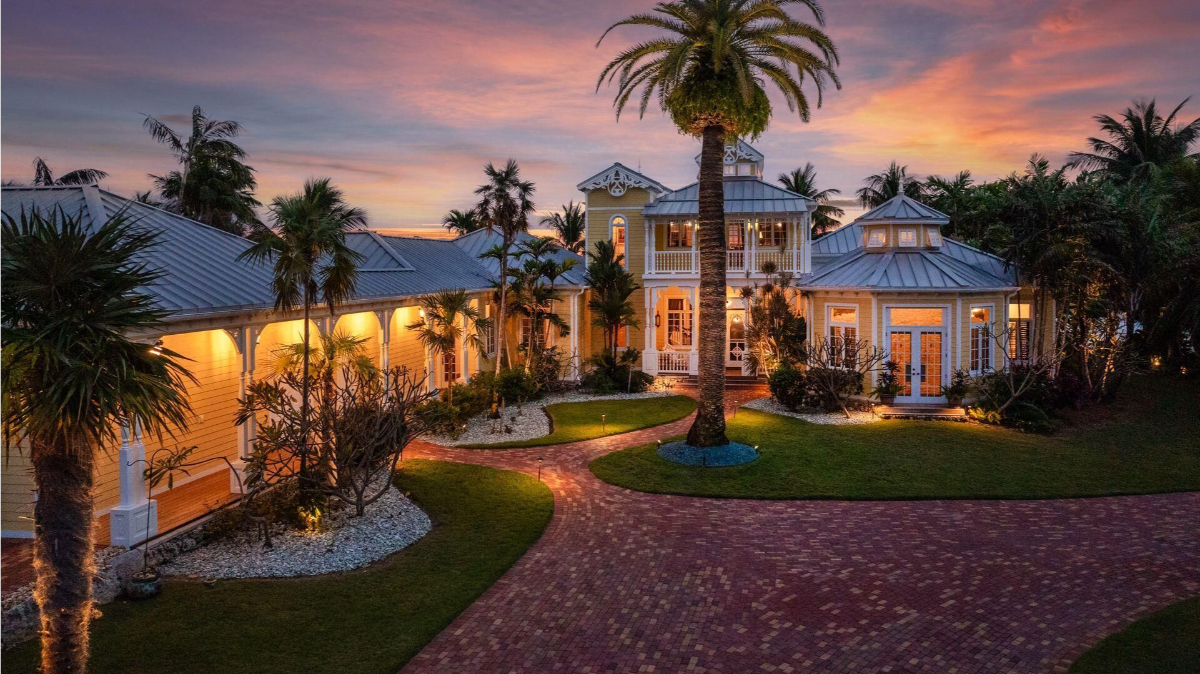 Coastal mansion exudes charm at twilight, framed by palm trees, glowing exterior lights, and a rich red brick driveway under a vibrant sunset sky.