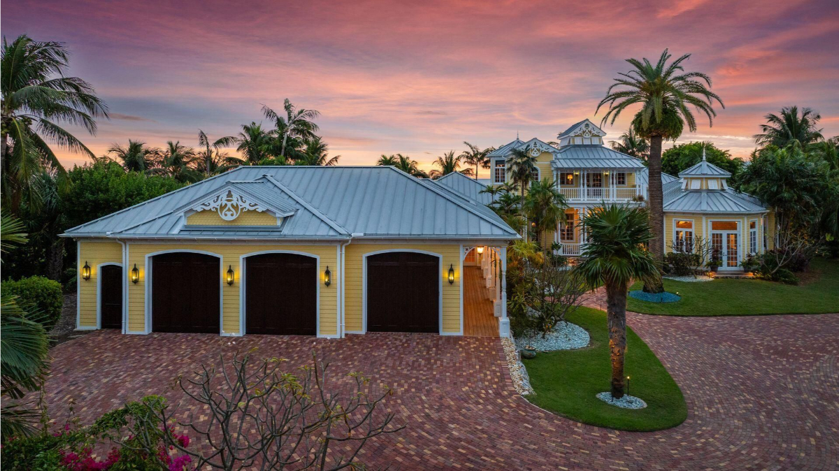 A pastel-hued sunset frames a coastal estate featuring a metal roof, warm lighting, and a brick driveway surrounded by lush tropical landscaping.