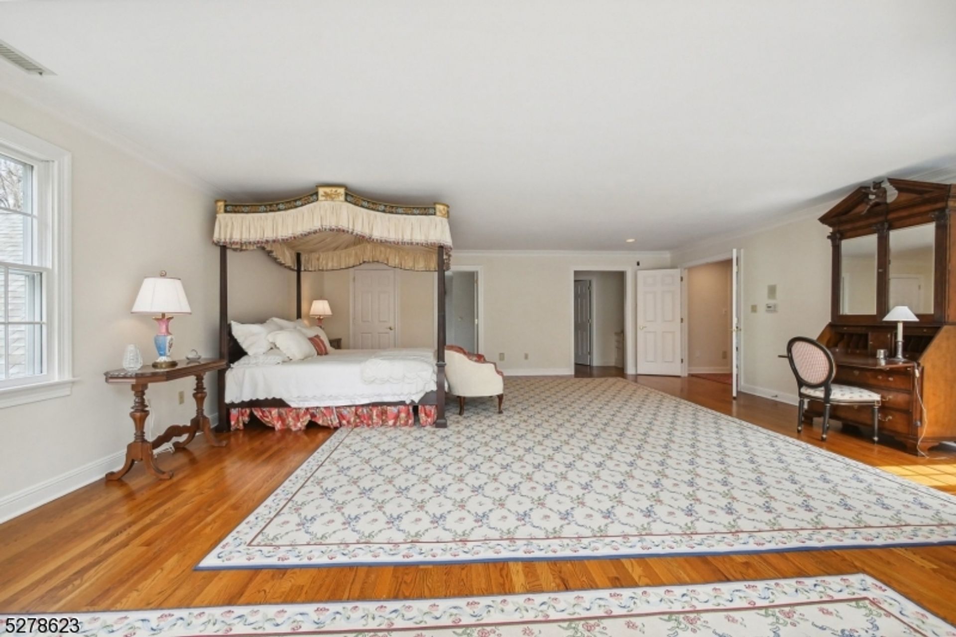 Spacious primary suite highlights a grand canopy bed, elegant writing desk, and floral-patterned area rugs over gleaming hardwood floors.