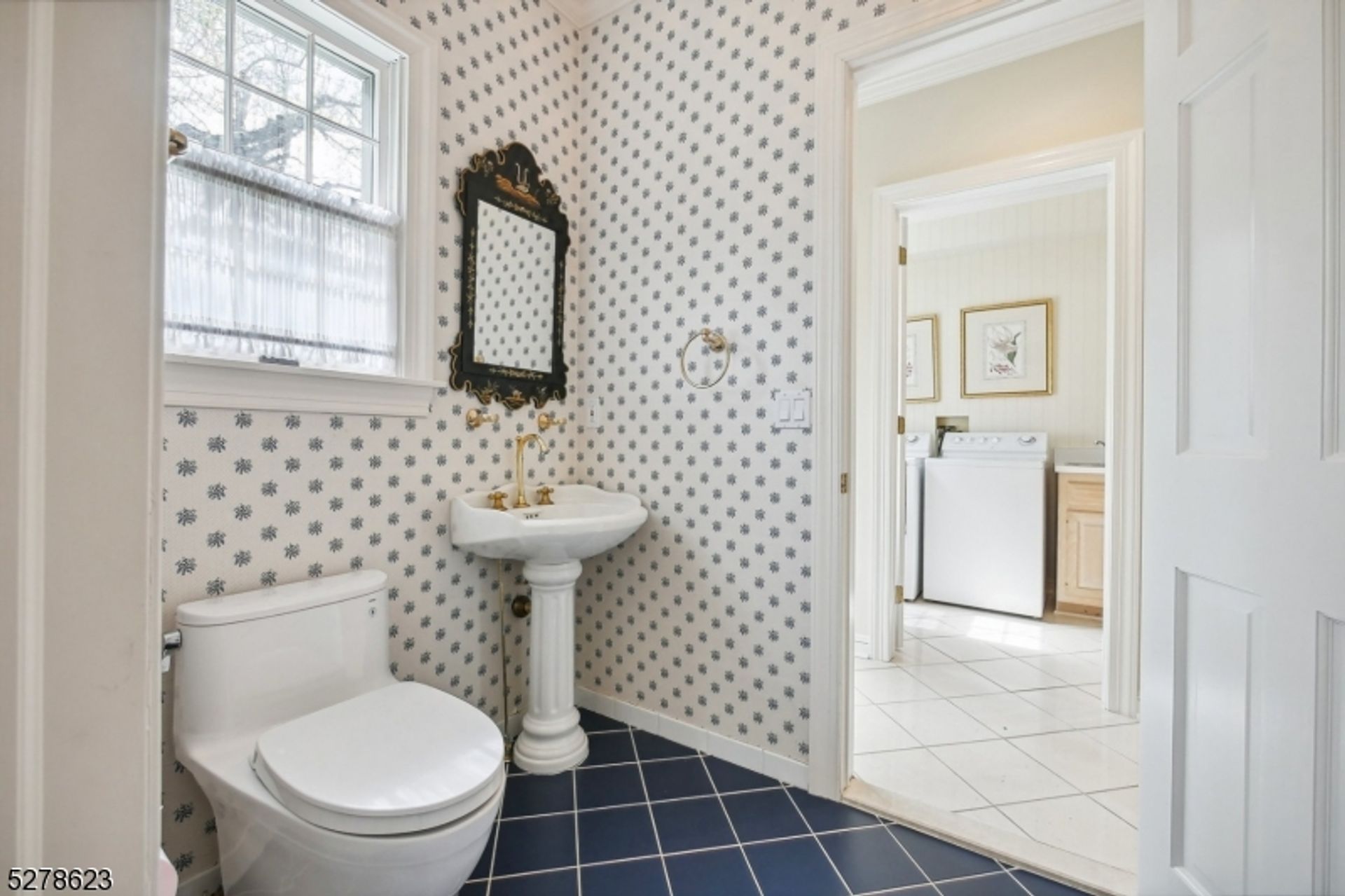 Powder room features patterned wallpaper, a pedestal sink with gold fixtures, and contrasting navy tile flooring.