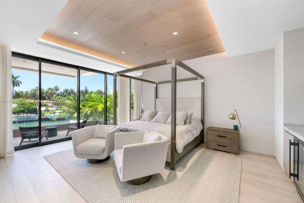 Modern bedroom with a canopy bed, cozy seating area, and floor-to-ceiling windows overlooking tropical views.