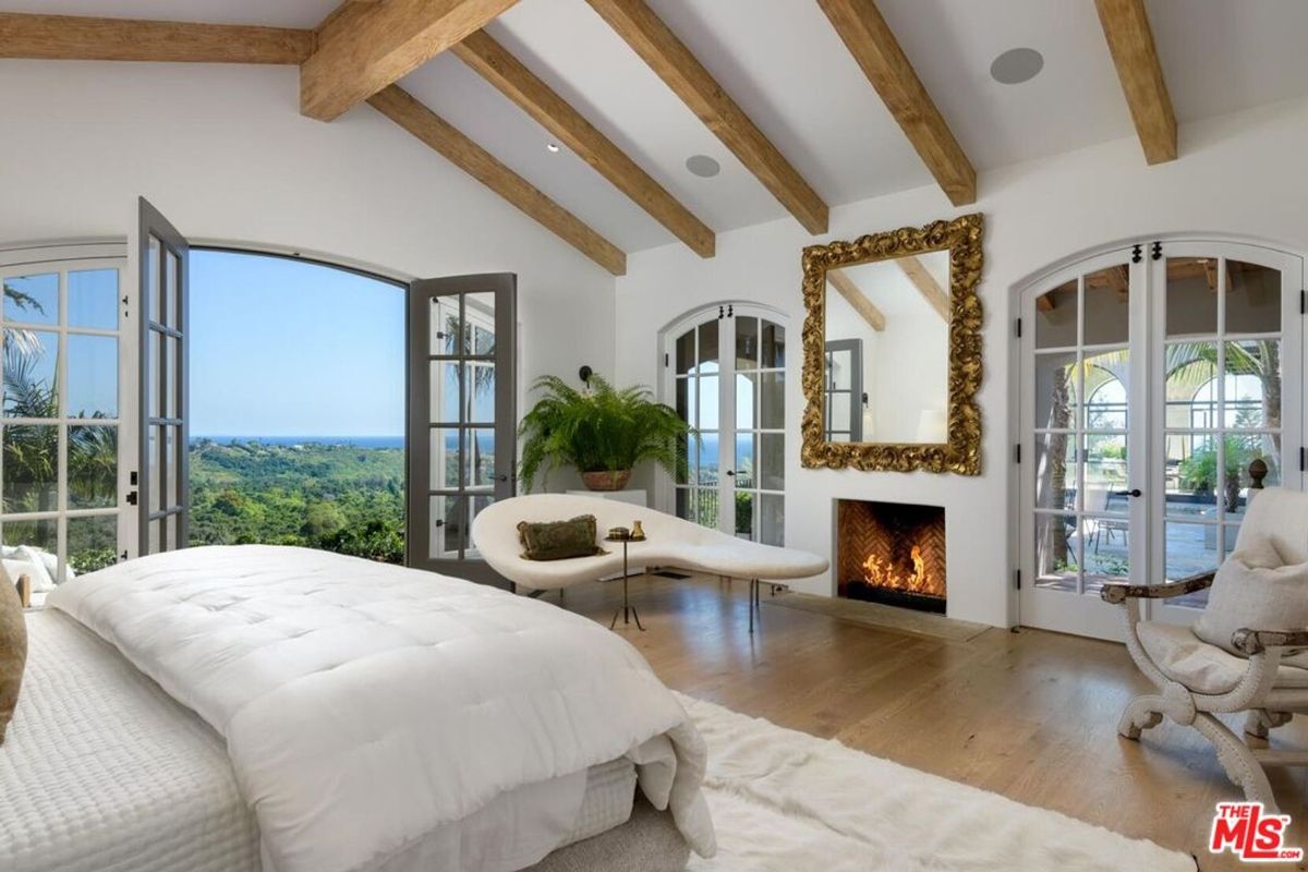 Vaulted ceilings with exposed wooden beams crown this serene bedroom, where French doors frame breathtaking ocean views and a cozy fireplace anchors the luxurious space.