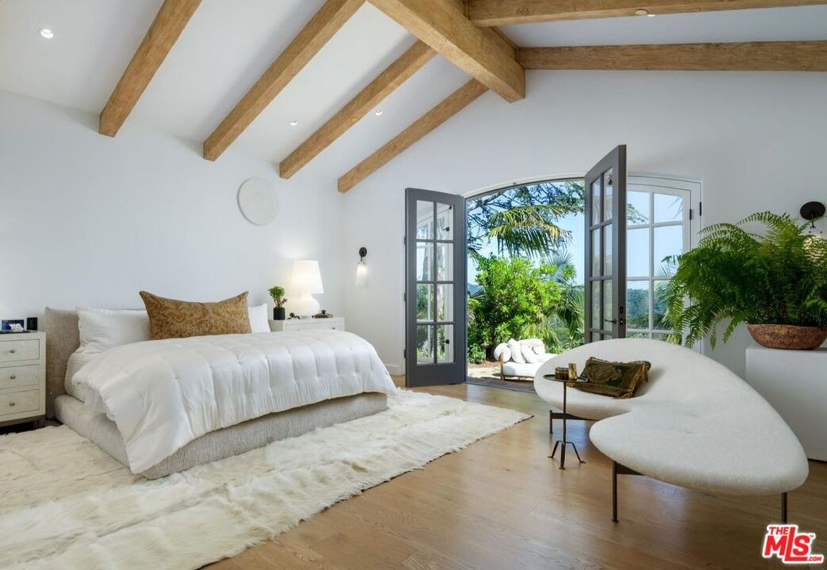 Exposed wood beams and natural light complement this tranquil bedroom, with French doors leading to lush greenery and a serene outdoor retreat.