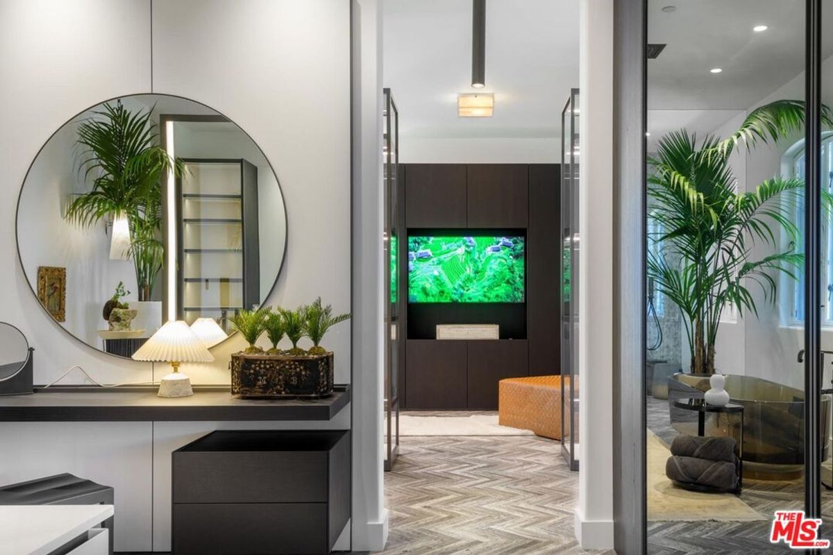 Sleek dressing area with a round mirror and lush greenery leads to a cozy lounge space with built-in cabinets and a vibrant TV display.