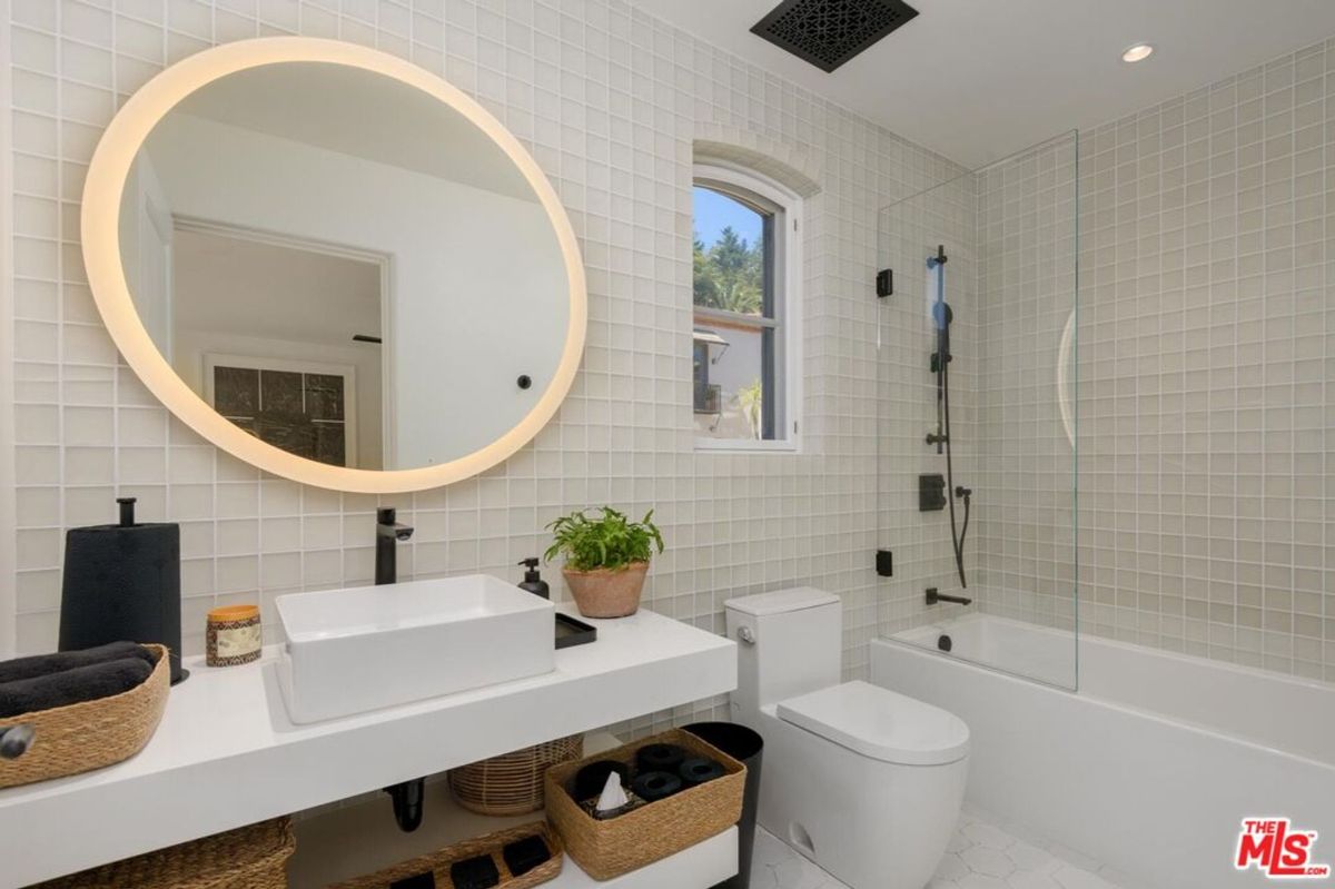 A chic bathroom with textured tile walls, a glowing round mirror, and modern black fixtures combines functionality with minimalist elegance.