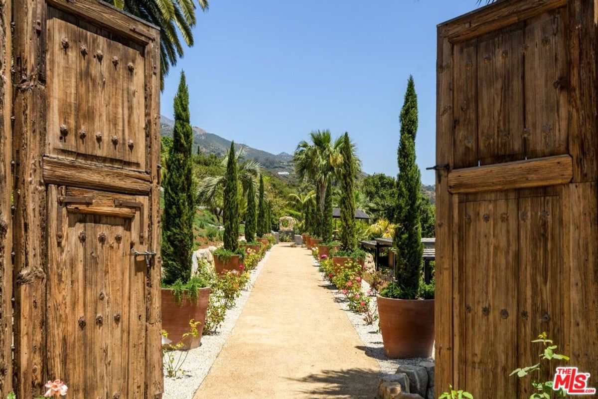 Gates reveal a serene pathway lined with towering cypress trees and lush greenery.