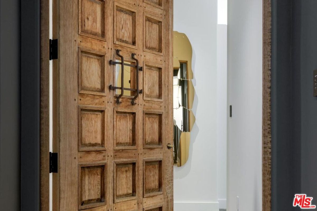 A rustic wooden door with intricate paneling opens into a modern interior featuring a unique gold-edged mirror.