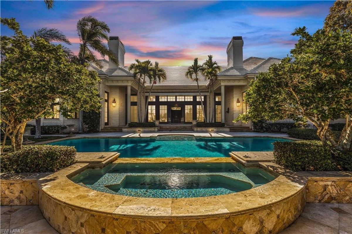 A luxurious pool area featuring a spa with stunning stonework is framed by lush landscaping and illuminated by a captivating sunset backdrop.