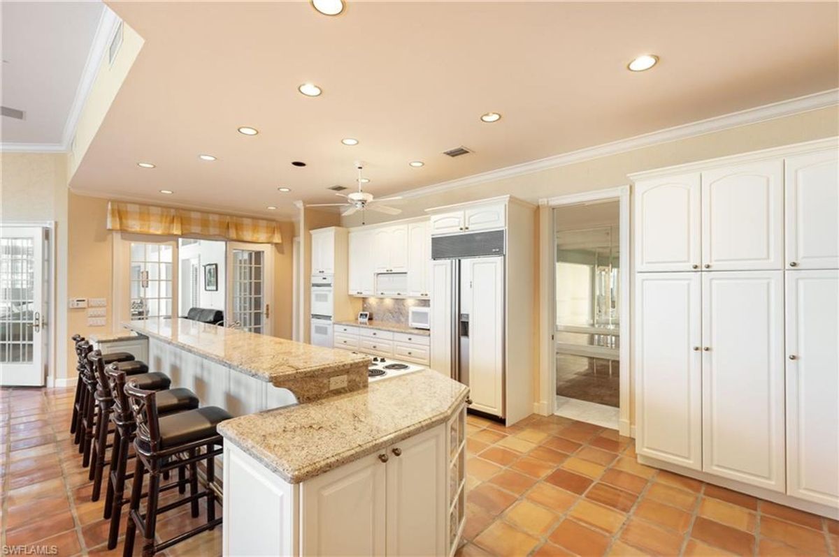 A gourmet kitchen boasts a long granite island with bar seating, white cabinetry, and integrated appliances, all complemented by warm terracotta flooring.