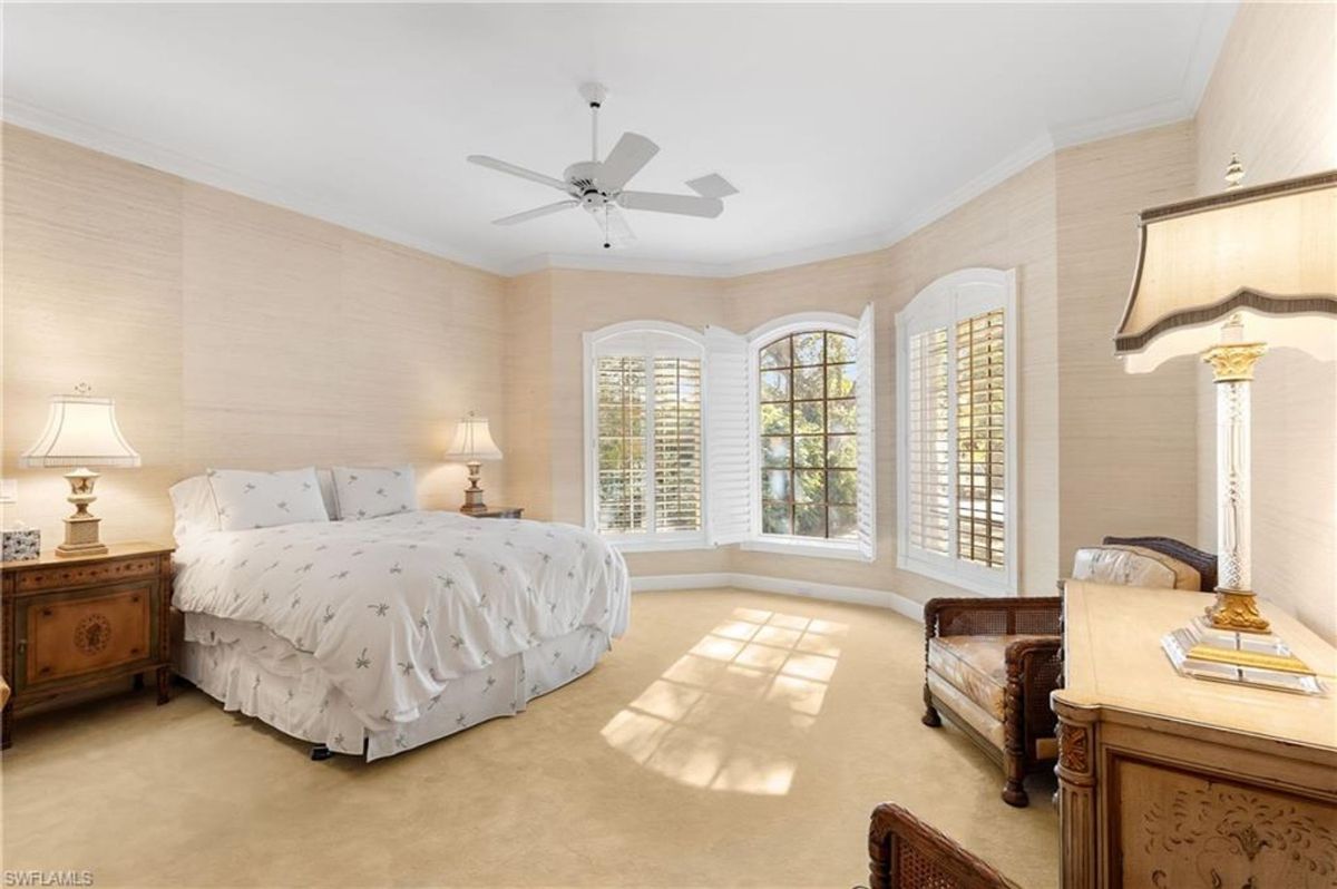 Bedroom features a cozy ambiance with neutral tones, bay windows, and elegant shuttered panels, creating a serene retreat.