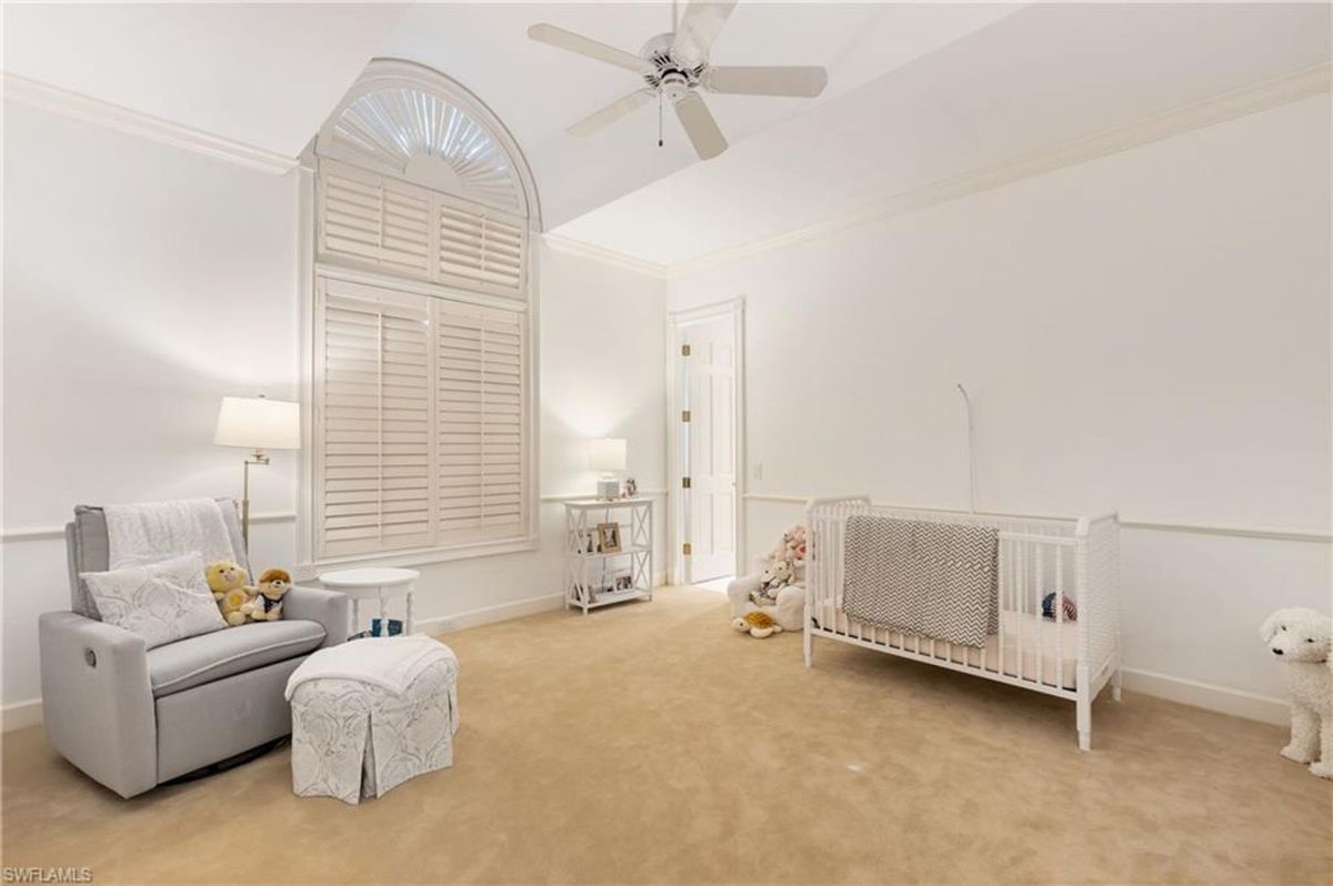 A charming nursery offers a cozy armchair, a crib, and soft neutral tones accentuated by an arched window with plantation shutters.