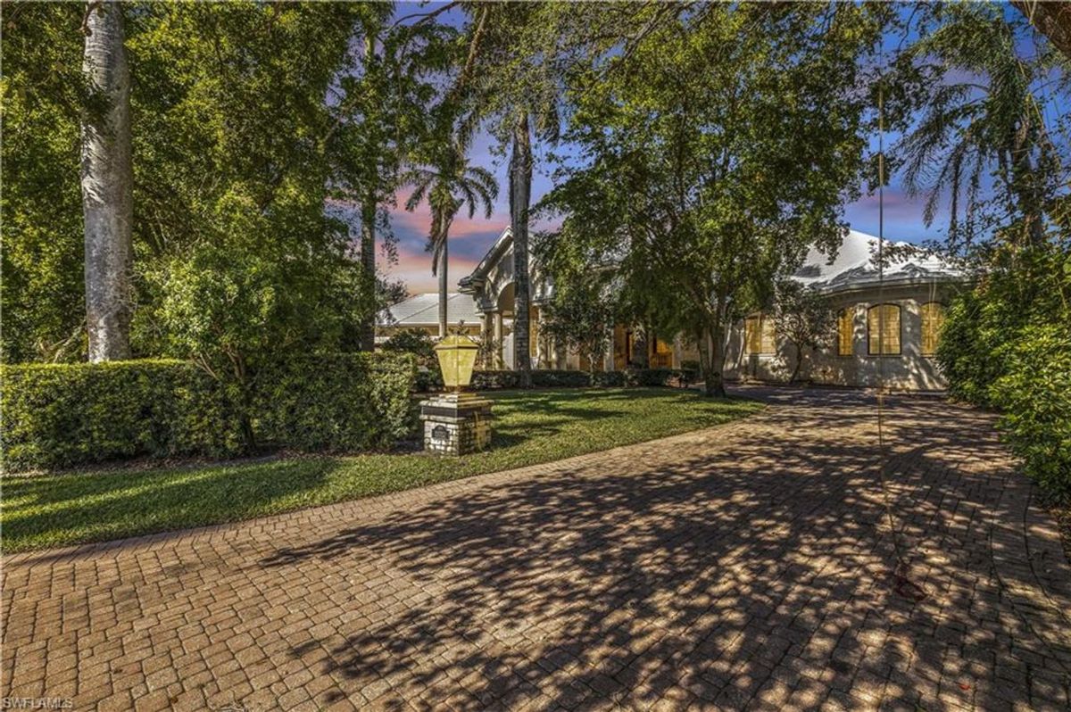 Driveway lined with lush greenery and tall palm trees leads to an elegant estate framed by soft evening hues in the sky.