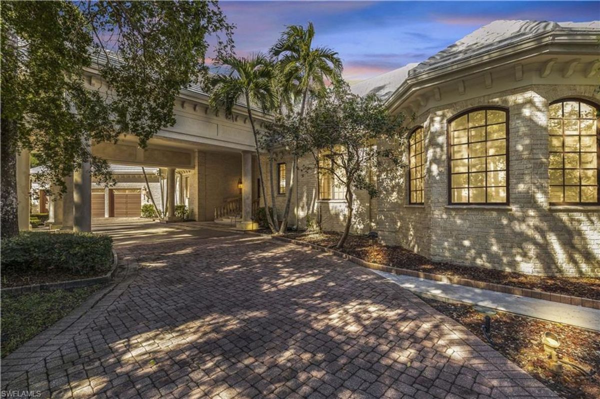 Brick-paved driveway shaded by mature trees leads to an elegant porte-cochère, blending timeless architecture with lush greenery.