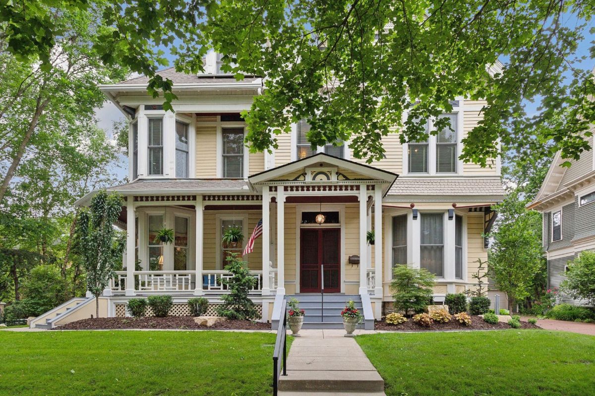 Large yellow Victorian house with a wraparound porch sits under leafy trees.