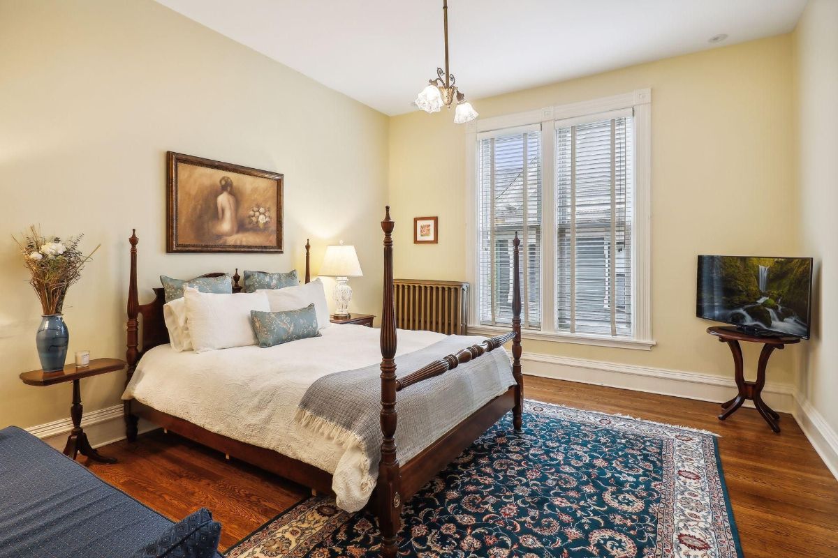 A bedroom with a four-poster bed, an area rug, and a large window is shown.