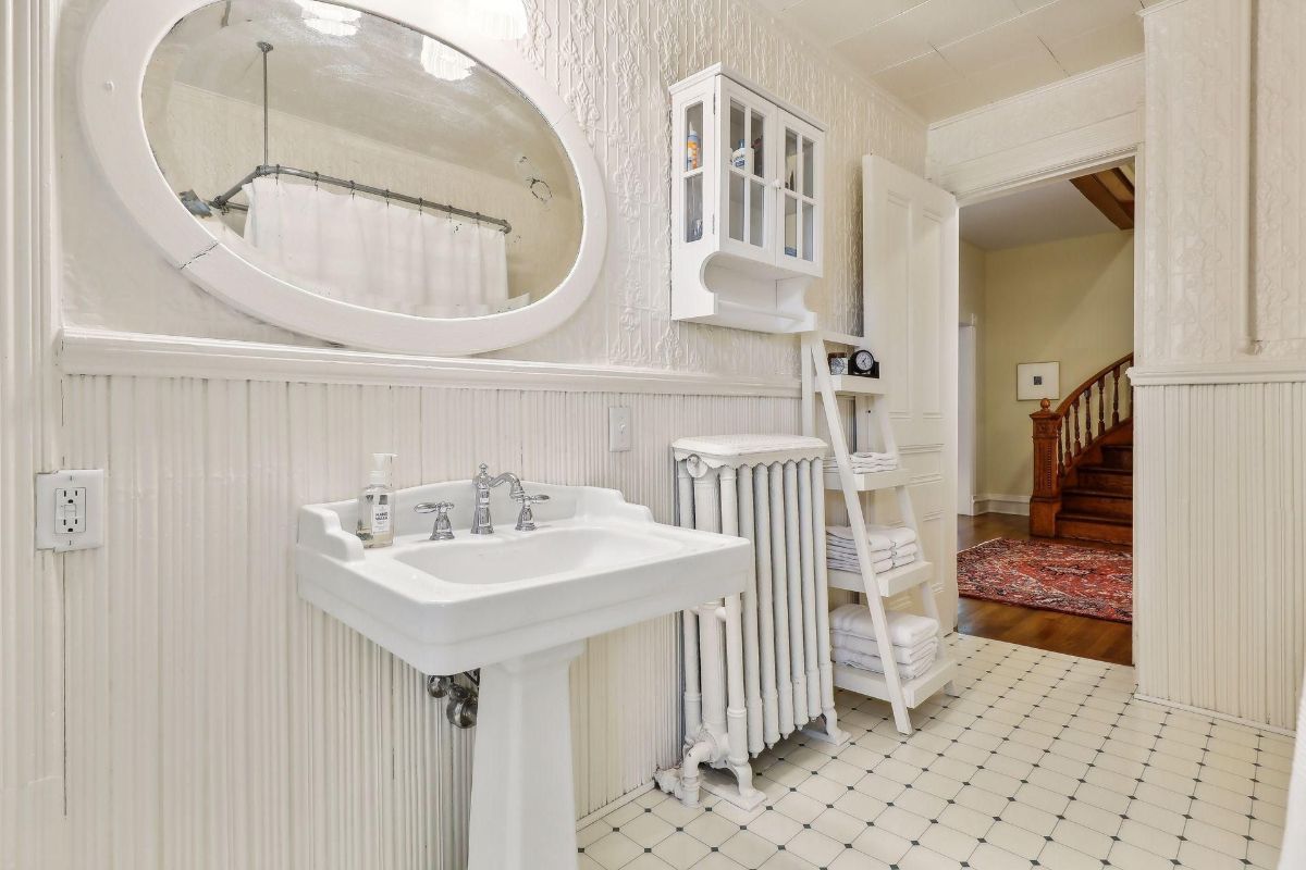 White bathroom with a pedestal sink, white tile floor, and a glimpse of a hallway.