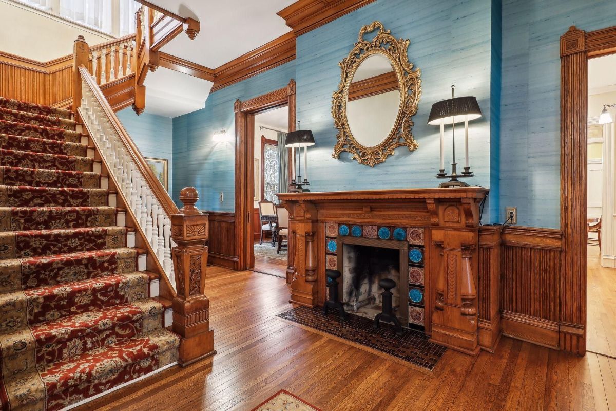 Grand staircase with a patterned runner is a prominent feature in this home's entryway, which also includes a fireplace with blue tile accents and a large mirror.