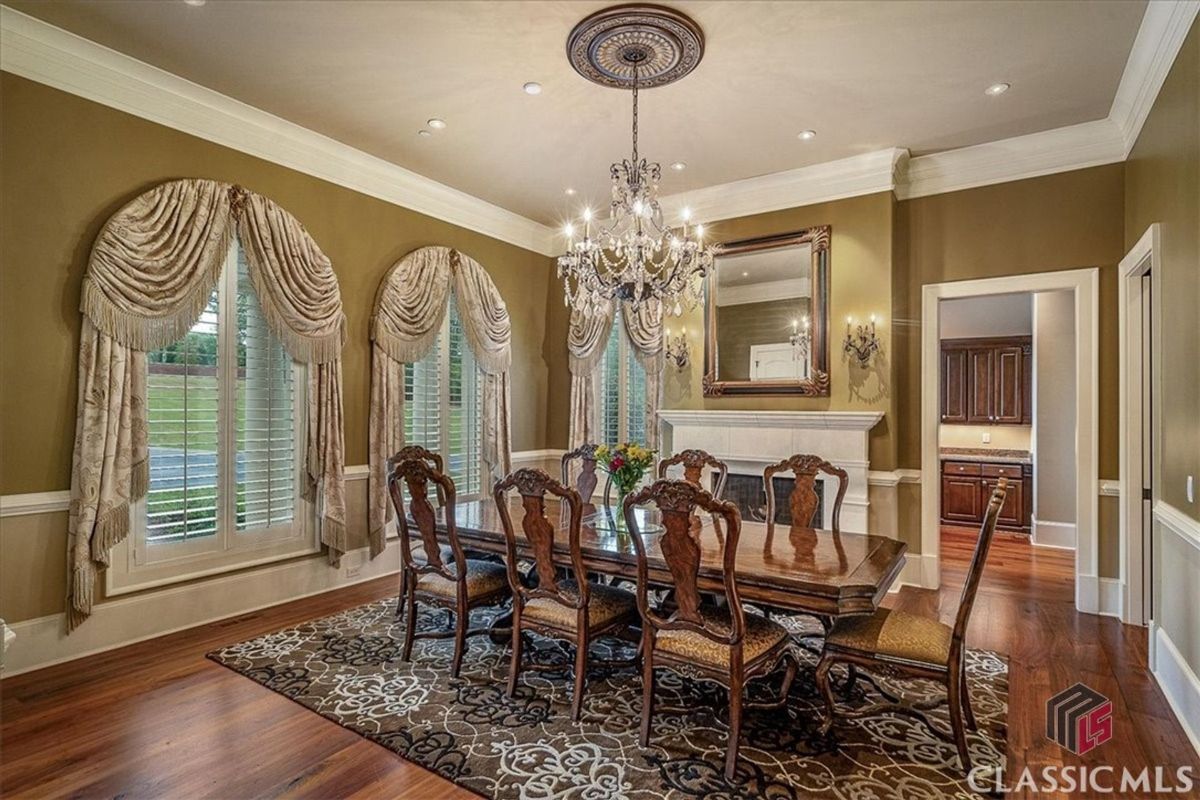 A formal dining area features a grand crystal chandelier, luxurious draped windows, a large ornate mirror above a classic mantle, and intricately designed chairs surrounding a polished wood table.