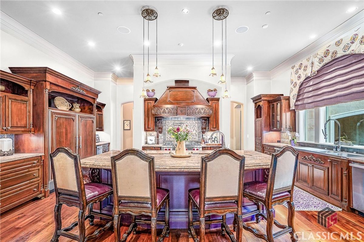 A gourmet kitchen showcases a large granite island with elegant high-back chairs, intricate wood cabinetry, custom range hood, and decorative pendant lighting.