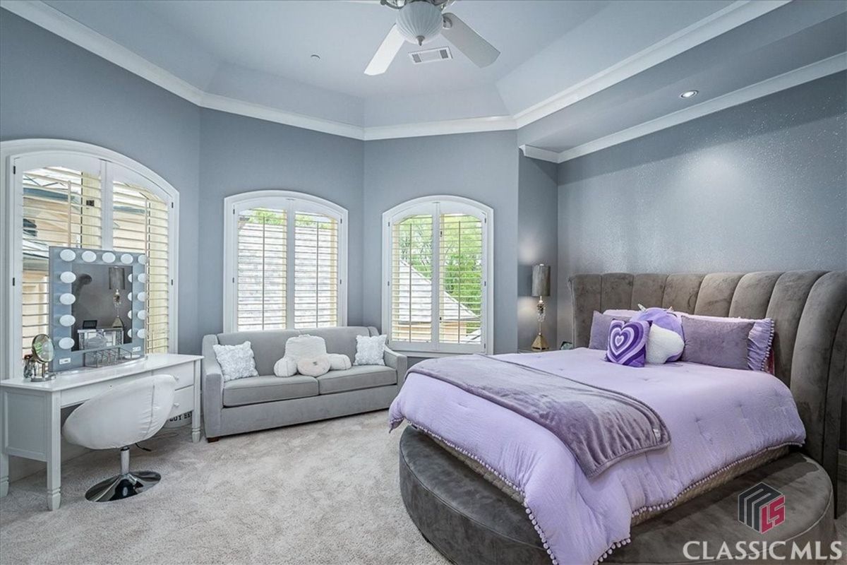 A serene bedroom offers soft lavender accents, a cozy sitting area, and abundant natural light through elegant arched windows.