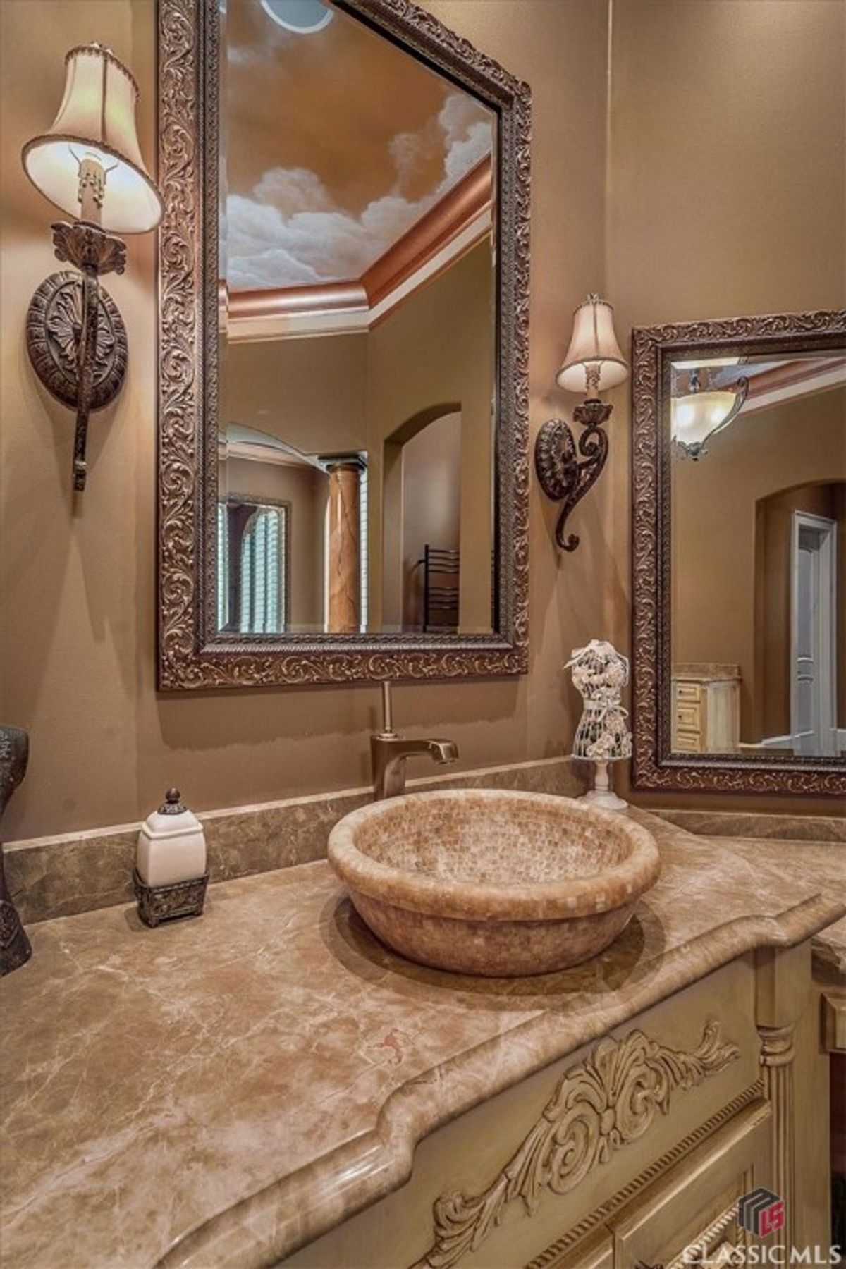 A luxurious bathroom detail featuring a marble vessel sink, intricate cabinetry, and a framed mirror reflecting the tray ceiling's cloud mural.