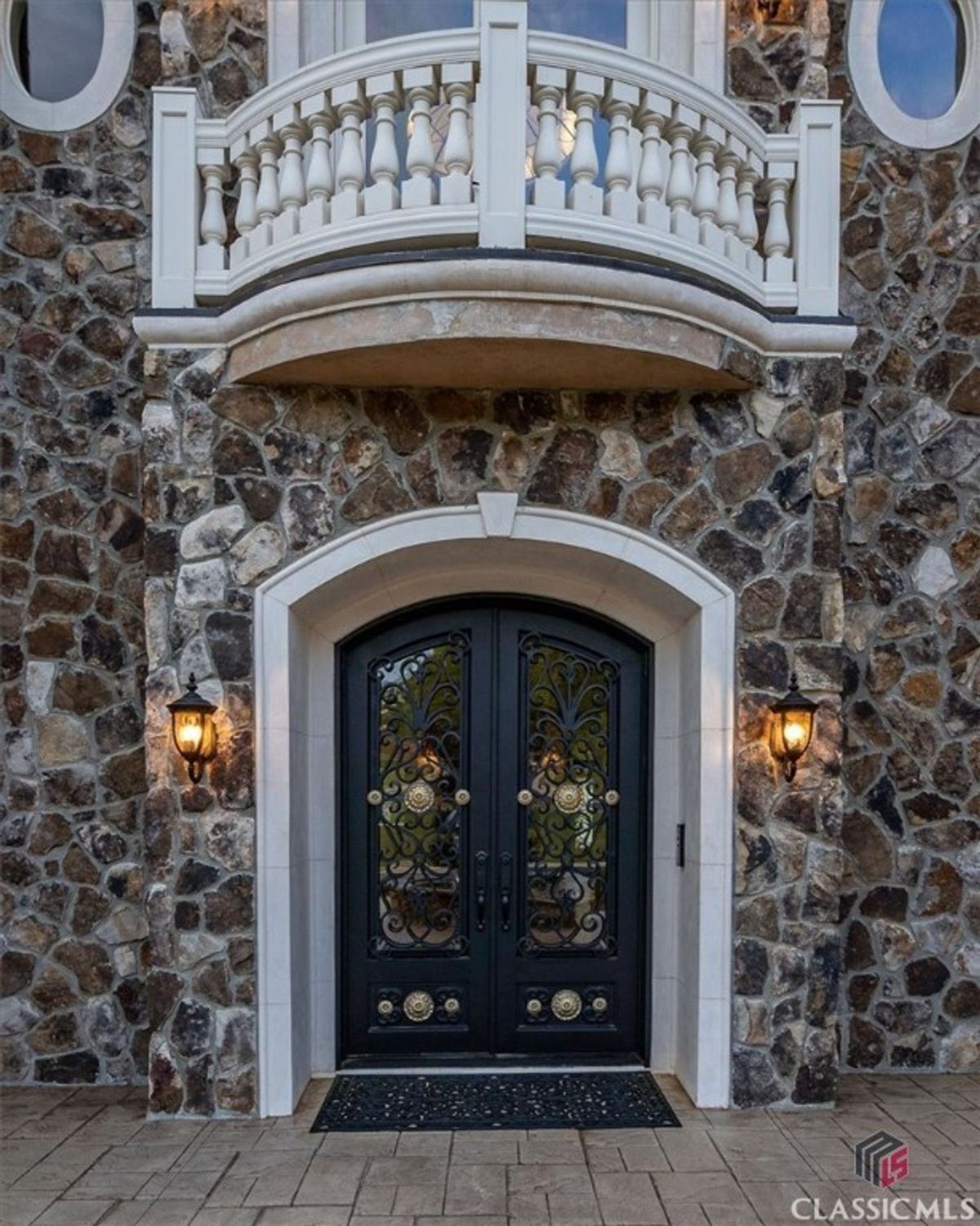 A grand iron double door framed by a rustic stone facade and topped with a charming Juliet balcony sets an inviting entrance.