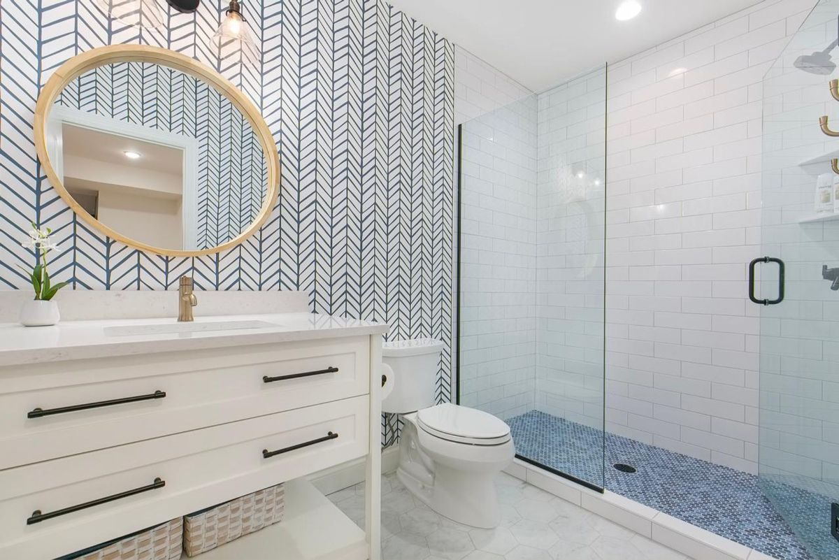 Stylish bathroom features a bold herringbone accent wall, a sleek glass shower, and a modern vanity with a round wood-framed mirror.