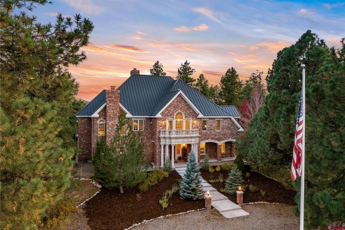 A large, multi-story house of brick and stone construction is shown. It features a dark-colored metal roof. Landscaping includes evergreens and a walkway. An American flag is visible on a flagpole. The setting sun casts a warm glow on the scene.