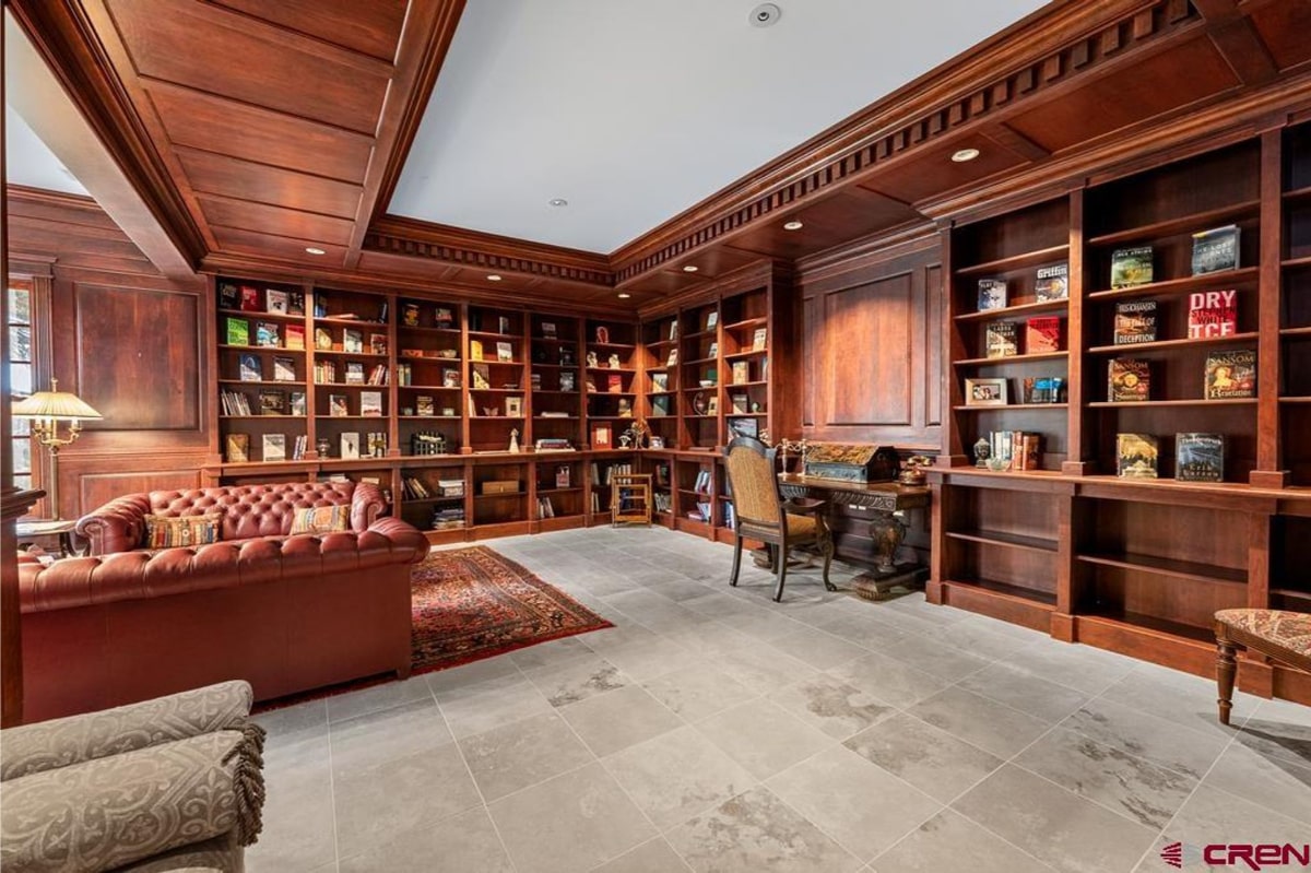 Luxurious library with rich, dark wood paneling and built-in bookshelves filled with books. A leather chesterfield sofa and a writing desk with a chair are also present in the room. The floor is light-colored stone tile, and an oriental rug sits in front of the sofa. The overall style is traditional and elegant.