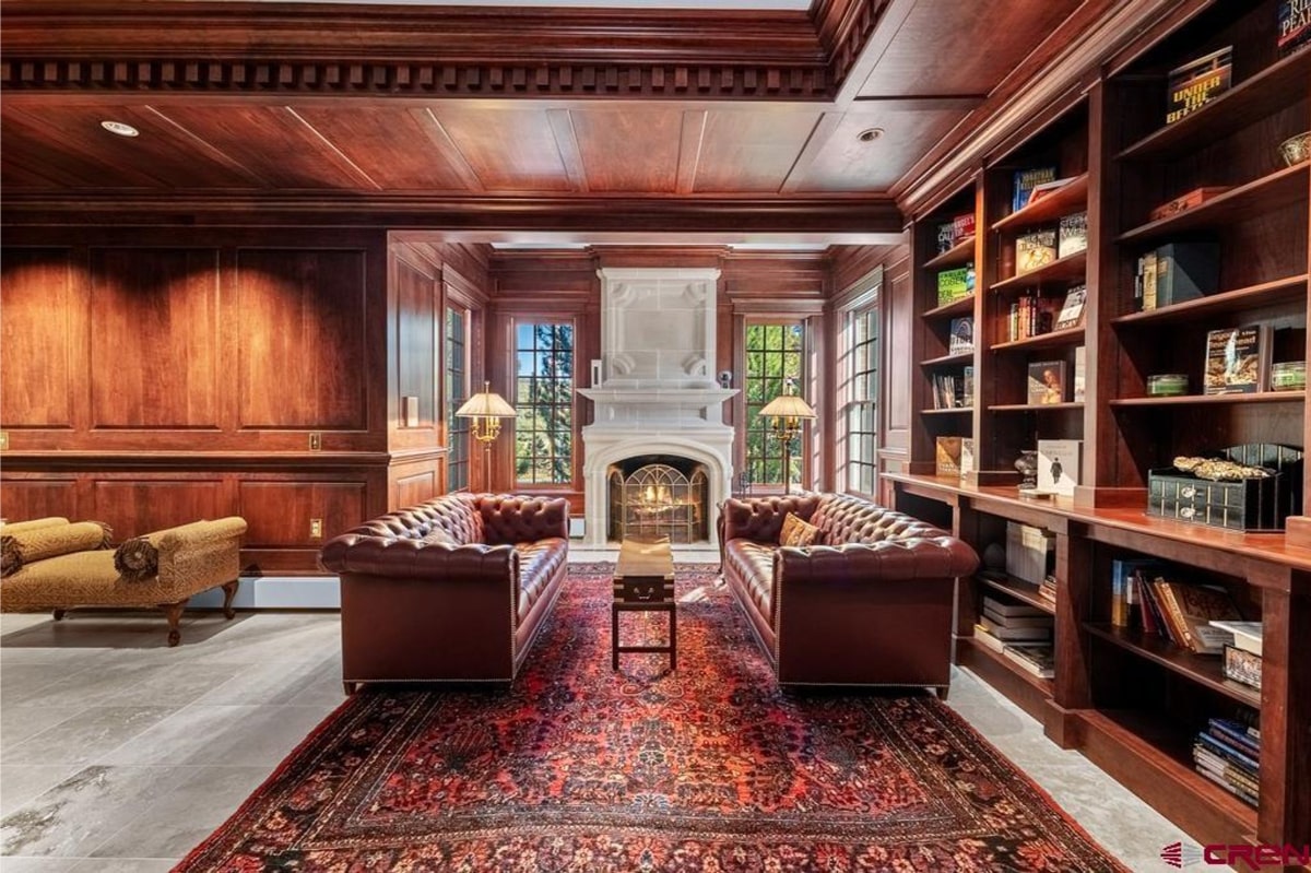 Luxurious living room or study with dark wood paneling on the walls and ceiling. Two large, brown leather chesterfield sofas face a large, ornate marble fireplace. There is a built-in bookcase in the corner of the room. An oriental rug covers the floor. The overall style is traditional and elegant.