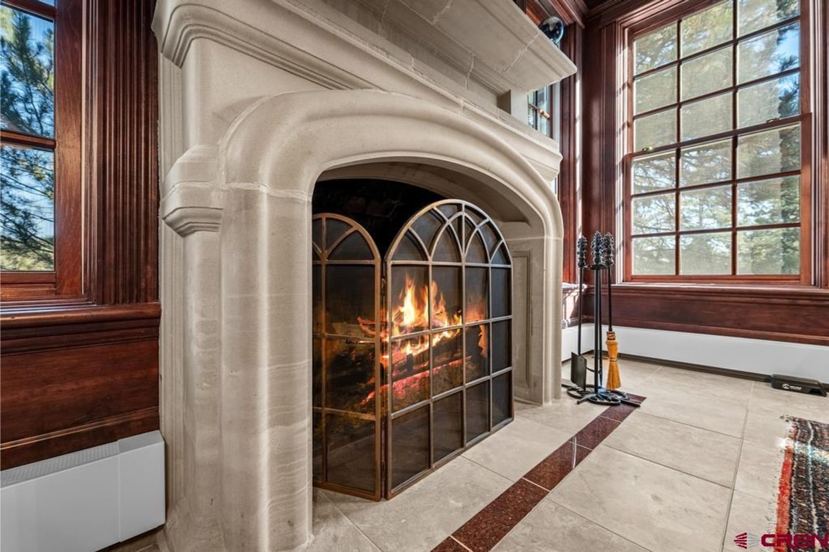 Large, ornate fireplace with a light-colored stone mantel and a metal fire screen. The fireplace is set into a wall with dark wood paneling, and a large window is visible in the background. A fireplace tool set is leaning against the wall next to the fireplace. The overall style is traditional and elegant.