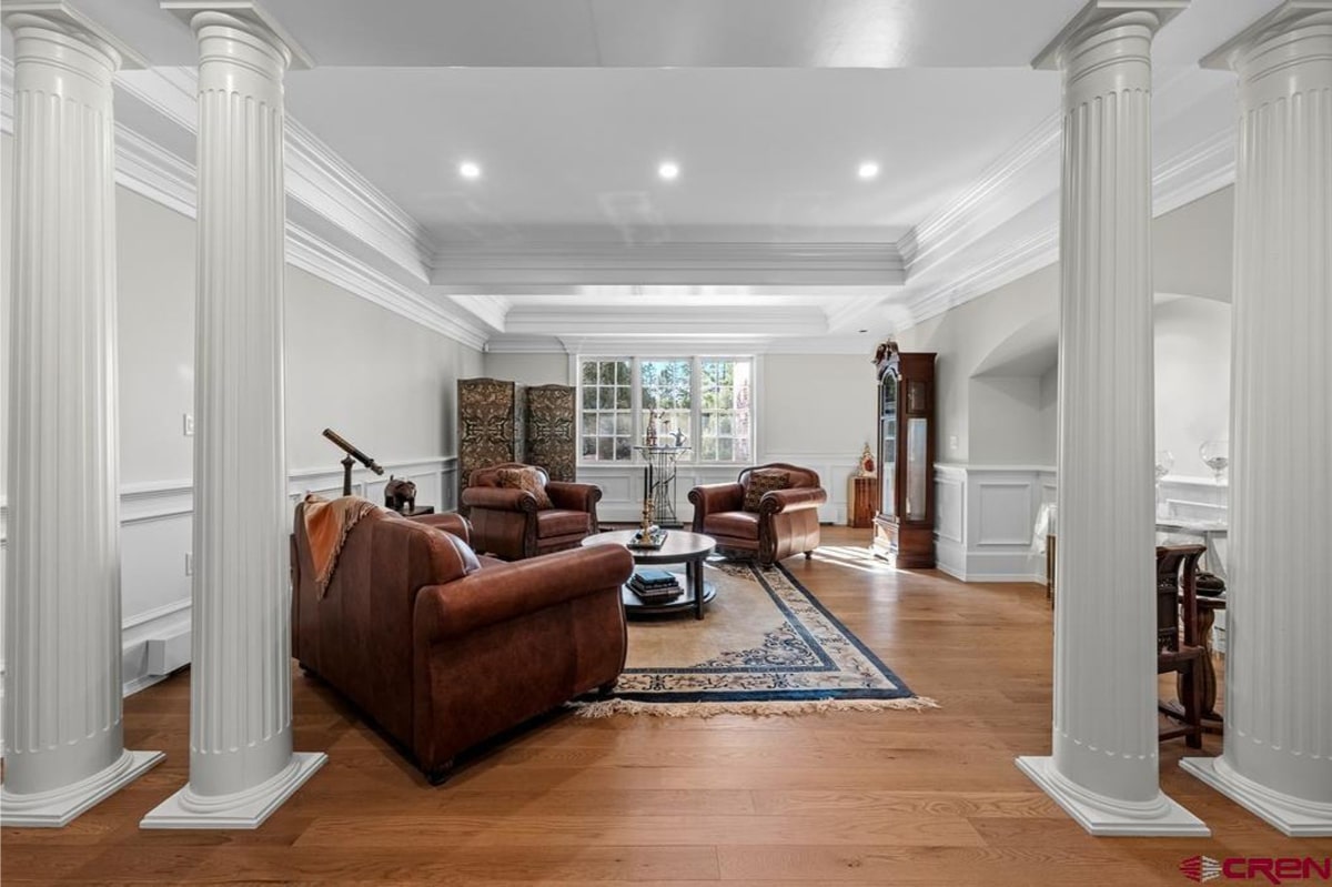 Spacious, elegantly furnished room with hardwood floors and white walls and trim. There are several brown leather armchairs and a sofa arranged around a coffee table on an area rug. A tall grandfather clock stands in the corner, and a telescope is visible on a stand near one wall. Large windows provide natural light. The overall style is sophisticated and traditional.