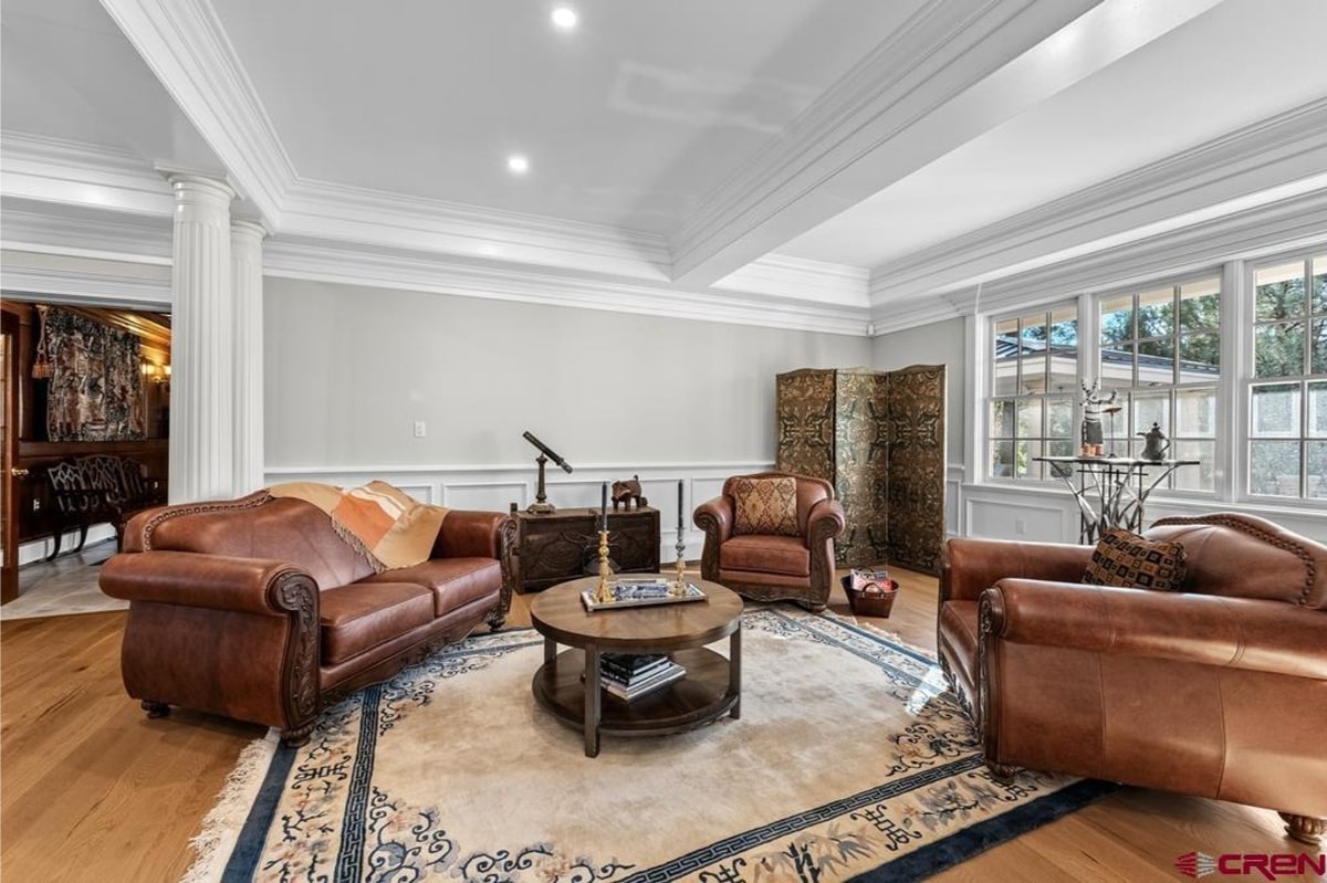 Luxurious living room with light-colored walls and hardwood floors. The room is furnished with several pieces of brown leather furniture, including a sofa and armchairs, arranged around a large round coffee table. A decorative screen stands in one corner, and a telescope is visible on a side table. The overall style is elegant and traditional