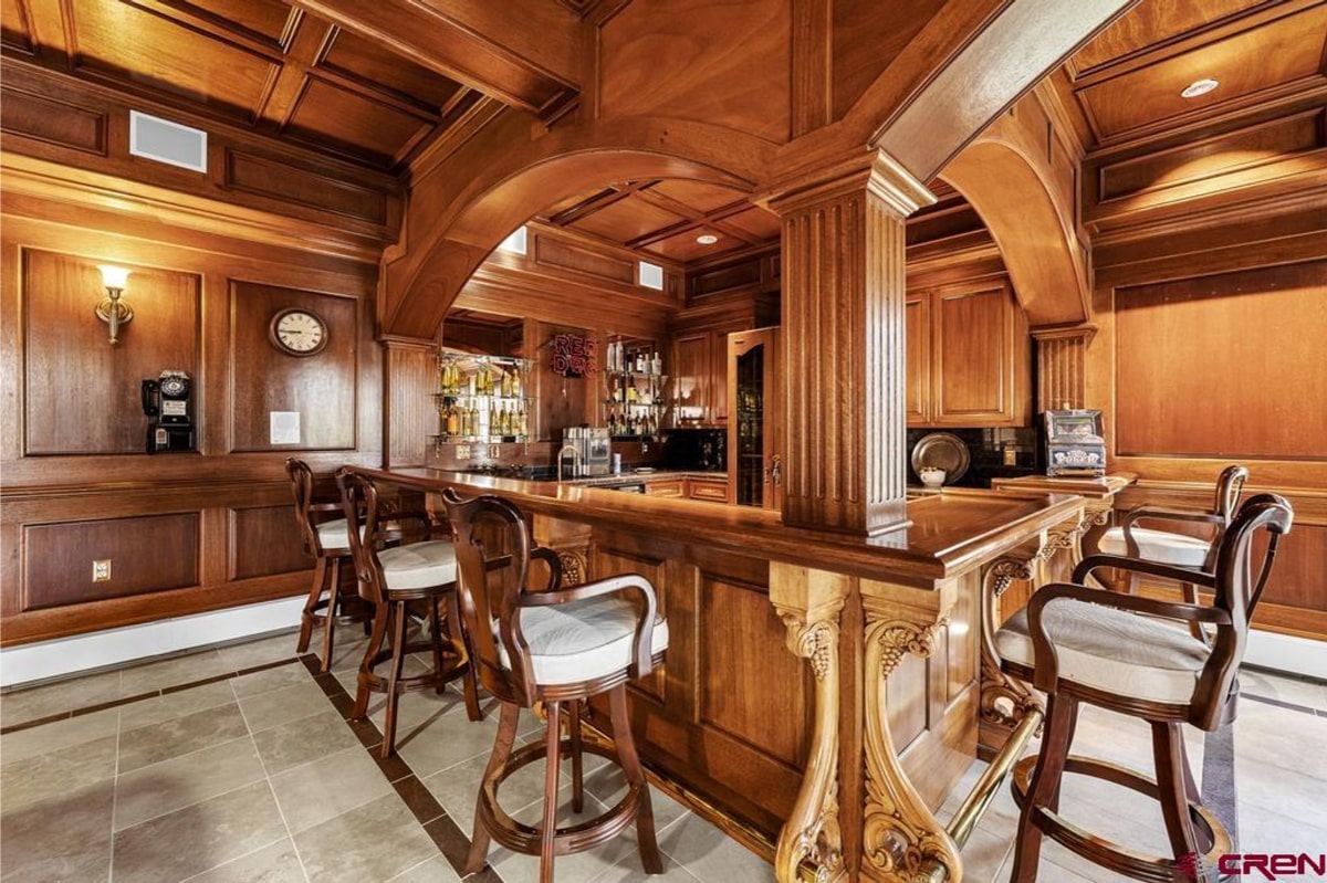 Luxurious home bar with rich, dark wood paneling and cabinetry. There's a long bar top with several high-top stools. The bar area has a traditional and elegant design. A vintage rotary phone is visible on the wall.