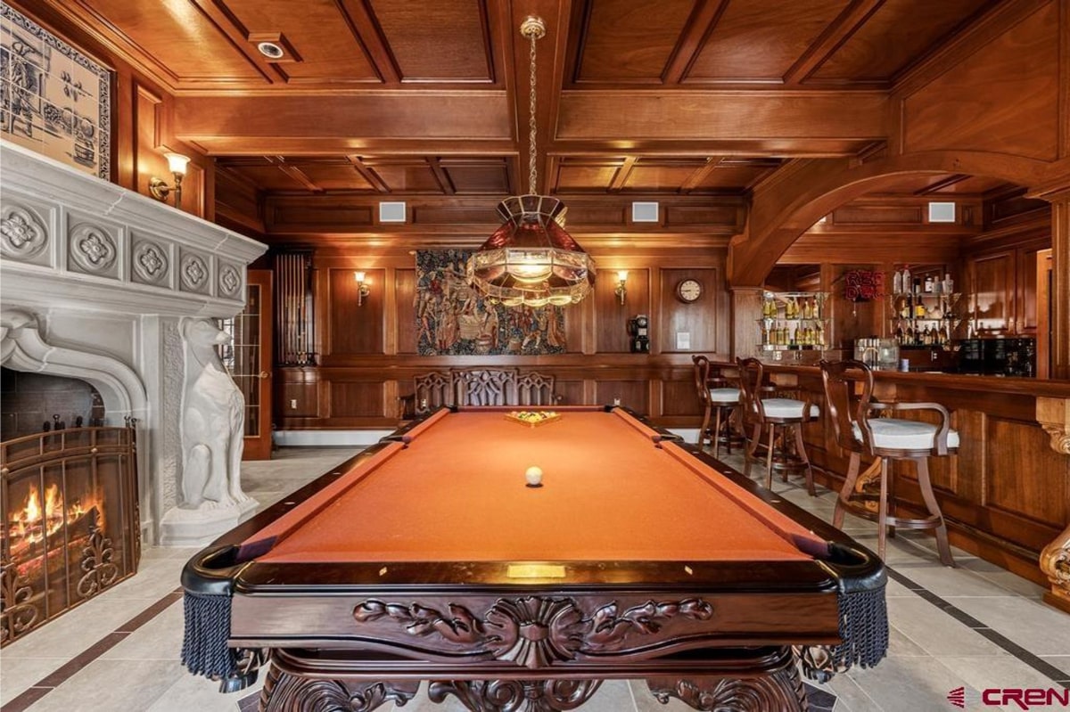 Luxurious game room with dark wood paneling, a large ornate pool table, and a bar area with high-top stools. There's also an elaborate fireplace and a tapestry on the wall. The overall style is traditional and opulent.