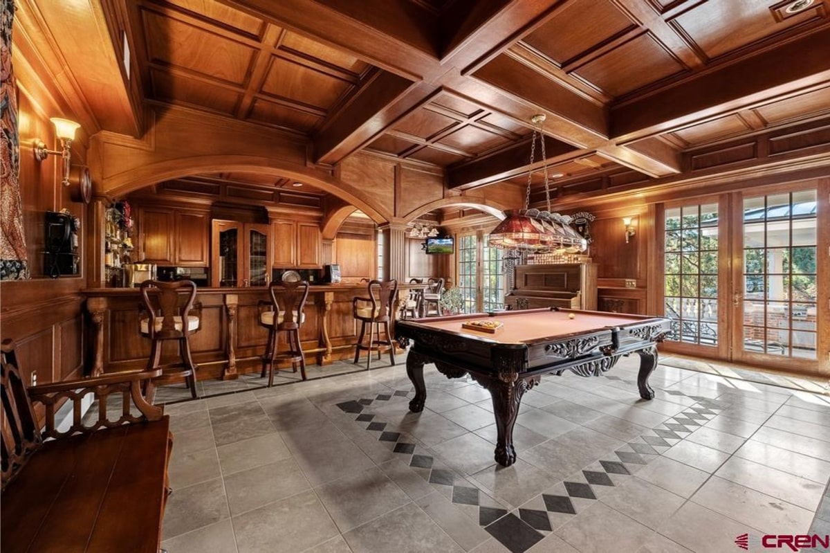 Game room with a pool table as the central feature. The room has a bar area with dark wood cabinetry and high-top stools. The walls and ceiling are also dark wood, creating a rich and traditional atmosphere. There are large windows that let in natural light. The floor is light-colored tile with a diamond pattern.