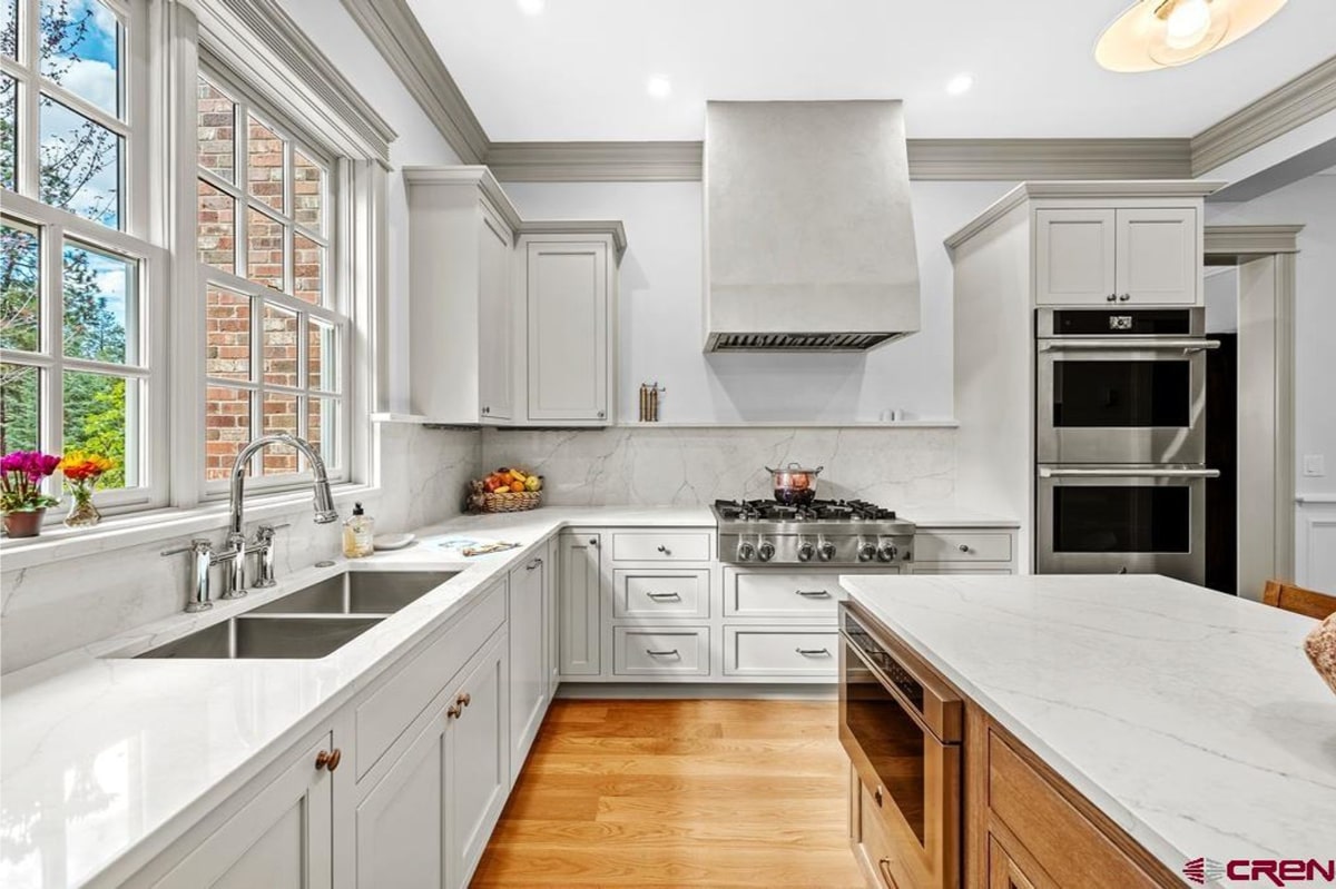 Bright, modern kitchen with white cabinets and countertops. There's a large island in the center of the room and stainless steel appliances. The backsplash is marble. A large window provides natural light. The floor appears to be hardwood. The overall style is clean and contemporary.